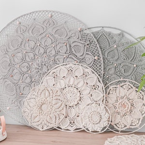 Op de afbeelding: Een verzameling handgemaakte, ronde wandkleden met ingewikkelde haakpatronen. De stukken variëren in grootte en kleur, van lichtgrijs tot crème, en zijn versierd met houten kralen. Een potplant is zichtbaar op de achtergrond.