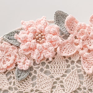 May include: A handmade crochet wall hanging featuring delicate pink flowers and gray leaves. The flowers have wooden bead centers. The background is a cream-colored, open-weave crochet pattern, creating a textured and artistic design.