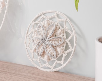Puede incluir: Un atrapasueños de crochet blanco con un diseño central gris. El atrapasueños está colgado en una pared blanca junto a una planta en maceta.