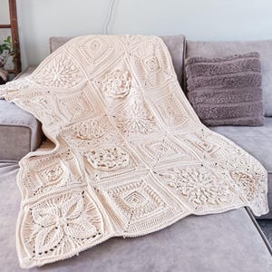 May include: A cream-coloured, handmade crochet blanket draped over a grey sofa. The blanket features a patchwork design with various floral and geometric patterns. A fluffy, grey cushion rests on the sofa. A cosy home decor item.