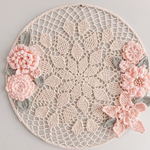 Op de afbeelding: Een gehaakt doily met een delicaat bloemenpatroon. De doily is crèmekleurig en heeft roze en grijze gehaakte bloemen.
