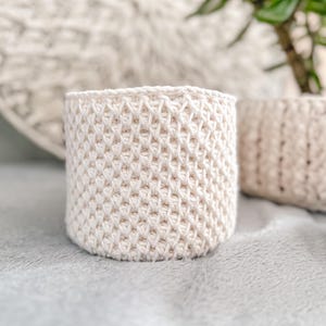 May include: A cream-coloured, cylindrical crochet basket with a textured, diamond-like pattern. The basket is made of thick yarn and is suitable for holding small items. Another similar basket is visible in the background.
