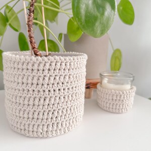 Può includere: Un vaso per piante all'uncinetto beige e una manica all'uncinetto più piccola per un barattolo di candele. Gli articoli fatti a mano sono esposti su una superficie bianca, con piante verdi e frondose sullo sfondo. Il vaso ha un diametro di circa 13 cm.