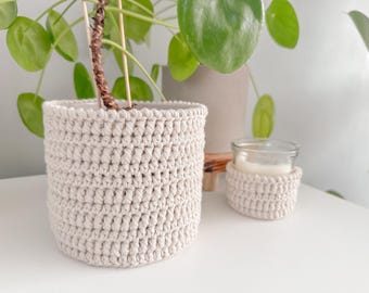 Puede incluir: Una maceta de ganchillo beige y una funda de ganchillo más pequeña para un tarro de velas. Los artículos hechos a mano se exhiben sobre una superficie blanca, con plantas de hojas verdes en el fondo. La maceta tiene unos 13 cm de diámetro.