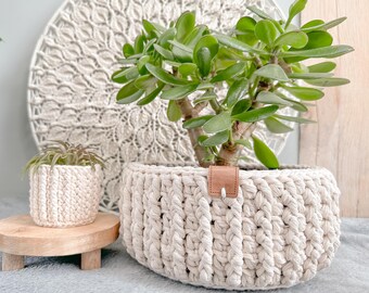 Puede incluir: Una maceta de crochet blanca con una correa de cuero marrón, que contiene una planta de jade verde. La maceta está sobre un soporte de madera.