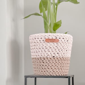 Op de afbeelding: Een roze en beige gebreide plantenpot met een bruin leren label waarop "Handmade with LOVE" staat.