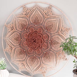 Op de afbeelding: Een grote, cirkelvormige wandhanger met een gedetailleerd, symmetrisch ontwerp. Het stuk is gemaakt van beige en bruine geweven materialen, waardoor een mandala-achtig patroon ontstaat. Het ontwerp heeft ingewikkelde details en een gradiënteffect, met een donkerder centrum. De wandhanger wordt tentoongesteld tegen een grijze muur.
