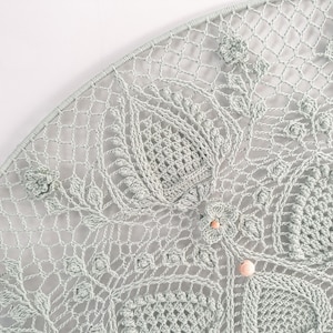 Puede incluir: Un tapiz de pared de crochet azul claro con forma circular y diseños florales intrincados. El tapiz presenta detalles delicados y una textura sutil.
