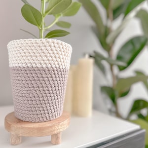 Puede incluir: Una pequeña planta en maceta en una maceta de ganchillo con la parte superior blanca y la inferior marrón claro. La maceta está sobre un soporte de madera con tres patas.