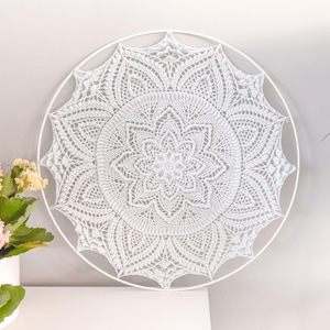 Op de afbeelding: Een witte ronde wandhanger met een ingewikkeld, met de hand gehaakt mandalapatroon dat strak in een bijpassende witte metalen ring is gespannen. Het ontwerp toont een symmetrisch, stervormig bloemmotief met gedetailleerd opengewerkt stikwerk.
