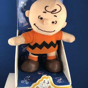 May include: A Charlie Brown plush doll in its original box. The doll has a peach-colored head with black hair and a smiling face. He wears an orange shirt with a black zig-zag pattern and brown shoes. The box features Snoopy and friends.