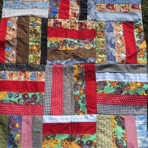 May include: A patchwork quilt constructed from various fabric strips featuring patterns of monster trucks, construction vehicles, red and white polka dots, black and white gingham, and solid red, brown, and green panels stitched together in a grid.