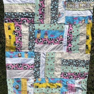 May include: A rectangular patchwork quilt constructed from various fabric strips featuring diverse patterns including soccer balls, blue hearts, yellow bees, mermaids, and green gingham. The quilt displays a multi-colored design with white, blue, pink, yellow, and green sections stitched together in a grid-like arrangement.