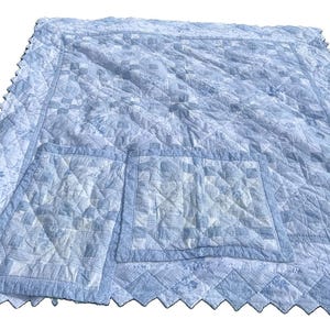 May include: A light blue and white quilted blanket featuring a patchwork design with floral and checkered patterns. The quilt has a scalloped edge and includes two square pockets stitched onto the surface, all in matching blue and white tones.