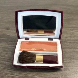 May include: Open compact of Elizabeth Arden blush with a mirror and brush. The blush is a peach color, and the brush has a dark brown handle with a gold ferrule. The compact is burgundy with a white interior.
