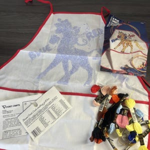 May include: A white fabric play smock with red trim featuring a blue stamped giraffe design and the text BLUE RED. Includes a Vogart Crafts instruction sheet, a product packaging card with a giraffe illustration, and assorted colorful embroidery floss.