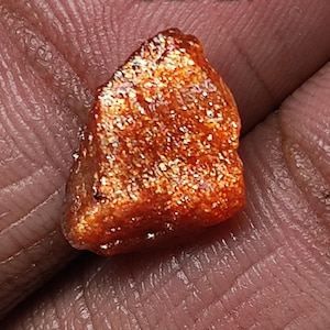 May include: A rough, orange-red sunstone with a sparkly, iridescent finish. The stone is irregularly shaped and has a slightly pointed tip.