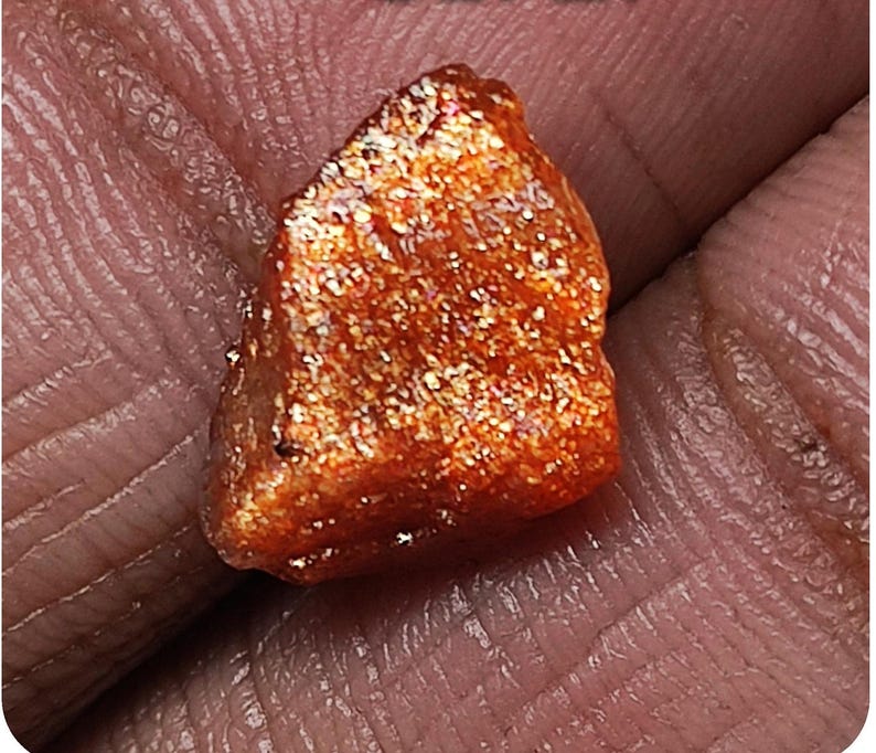 May include: A rough, orange-red sunstone with a sparkly, iridescent finish. The stone is irregularly shaped and has a slightly pointed tip.