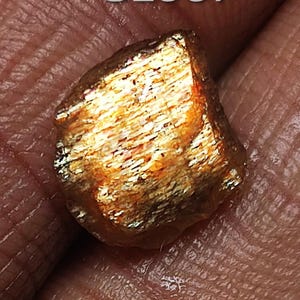 May include: A rough, orange-gold sunstone with a shimmering, iridescent surface. The stone is irregularly shaped and has a natural, unpolished finish.