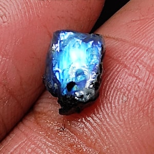 Puede incluir: Una piedra preciosa azul con una forma irregular y rugosa. La piedra preciosa tiene un brillo iridiscente y está engastada en una matriz gris oscuro.