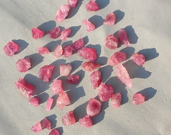May include: A collection of pink and red rough gemstones, likely rose quartz, scattered on a white surface. The stones are various sizes and shapes, some with sharp edges and others more rounded.
