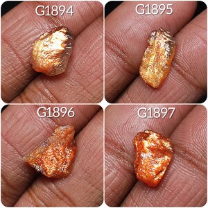 May include: Four sunstone specimens, each with a unique shape and color variation. The stones are a mix of orange, red, and gold, with a shimmering, iridescent effect. The stones are labeled with the numbers G1894, G1895, G1896, and G1897.