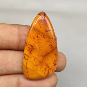 Puede incluir: Una piedra preciosa en cabujón en forma de lágrima con una base amarilla y naranja y vetas rojas y marrones. La piedra está pulida y tiene una superficie lisa.