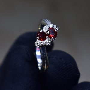 Può includere: Un anello d'argento con due pietre preziose rosse e pietre preziose trasparenti più piccole incastonate in una fascia curva.