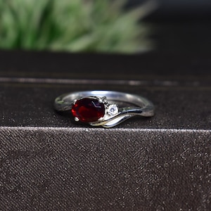 May include: A silver ring with a red oval gemstone and a smaller clear gemstone. The ring has a curved band.