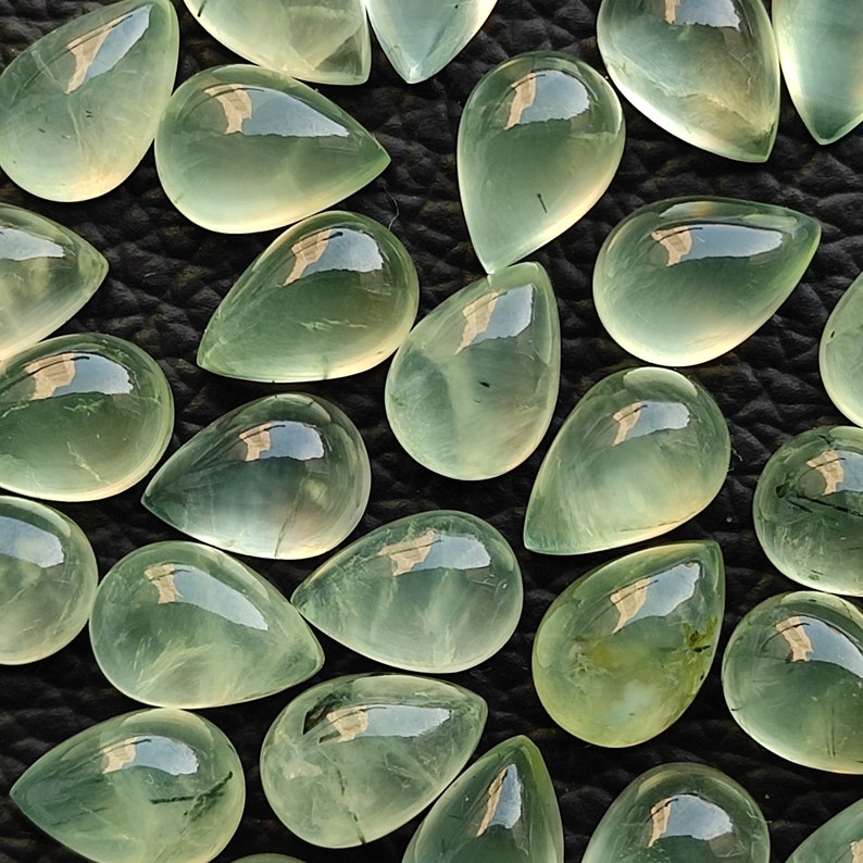 May include: A collection of green teardrop-shaped gemstones with a translucent appearance. The stones have a light green color and are arranged in a random pattern.