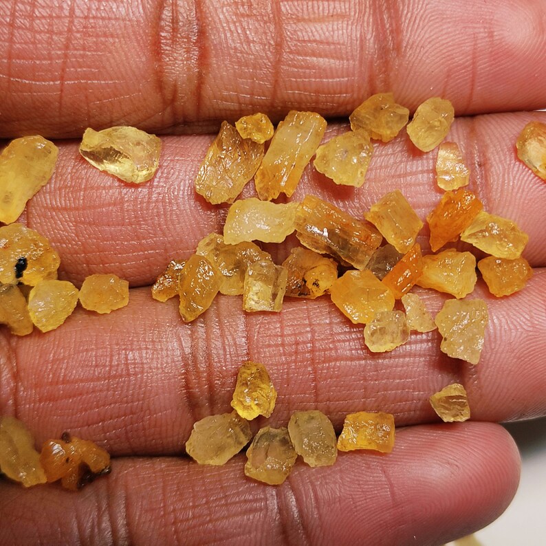 May include: A collection of small, rough, yellow-orange citrine crystals. The crystals are various shapes and sizes, and some have a translucent appearance.