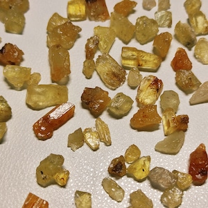 May include: A collection of small, translucent, yellow-brown crystals. The crystals are of various shapes and sizes, and some have a more orange hue.