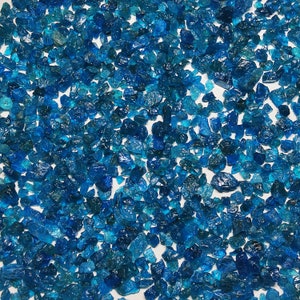 May include: A close-up image of a large quantity of blue apatite gemstones. The stones are various sizes and shapes, and they are scattered on a white surface.