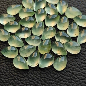 May include: A collection of teardrop-shaped green gemstones, also known as green aventurine, with a translucent, glassy finish. The stones are arranged in a cluster on a black surface.