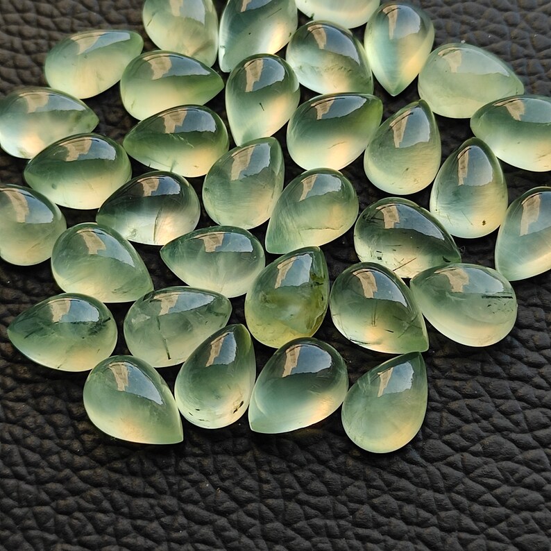 May include: A collection of teardrop-shaped green gemstones, also known as green aventurine, with a translucent, glassy finish. The stones are arranged in a cluster on a black surface.