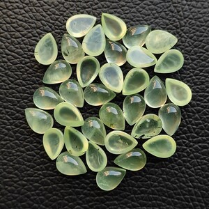 May include: A collection of 25 teardrop-shaped green gemstones with black inclusions. The stones are arranged in a loose pile on a black leather surface.