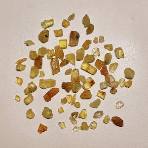 May include: A collection of small, yellow-green crystals, some with a more golden hue, scattered on a white surface. The crystals are of various shapes and sizes, with some appearing more translucent than others.