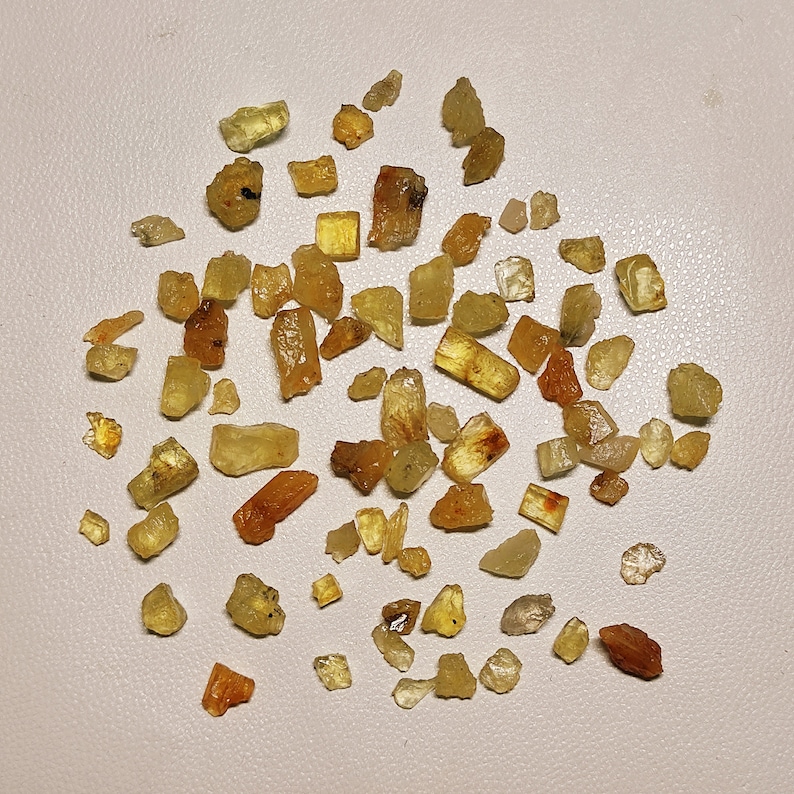 May include: A collection of small, yellow-green crystals, some with a more golden hue, scattered on a white surface. The crystals are of various shapes and sizes, with some appearing more translucent than others.