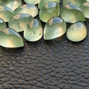 May include: A collection of green gemstone cabochons, each with a unique, pear-shaped cut. The stones have a translucent, glassy appearance with subtle inclusions.