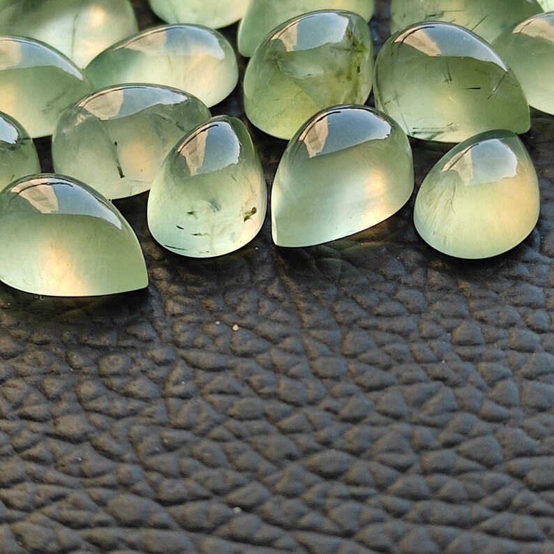 May include: A collection of green gemstone cabochons, each with a unique, pear-shaped cut. The stones have a translucent, glassy appearance with subtle inclusions.