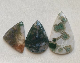 Op de afbeelding: Drie cabochons van mosagaat in een traanvorm. De stenen zijn een mix van wit, groen en bruin met een doorschijnende uitstraling.