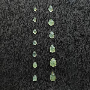May include: A collection of ten teardrop-shaped green gemstones of varying sizes arranged in two vertical rows on a black leather background.