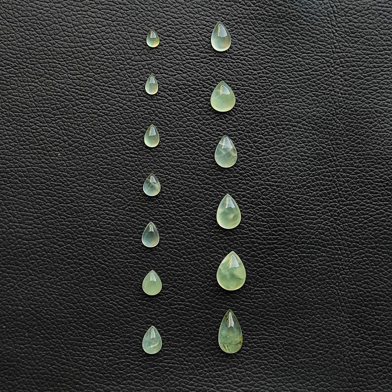 May include: A collection of ten teardrop-shaped green gemstones of varying sizes arranged in two vertical rows on a black leather background.
