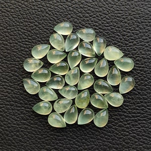 May include: A collection of 25 teardrop-shaped green gemstones. The stones are translucent and have a slight shimmer. They are arranged in a circular pattern on a dark background.