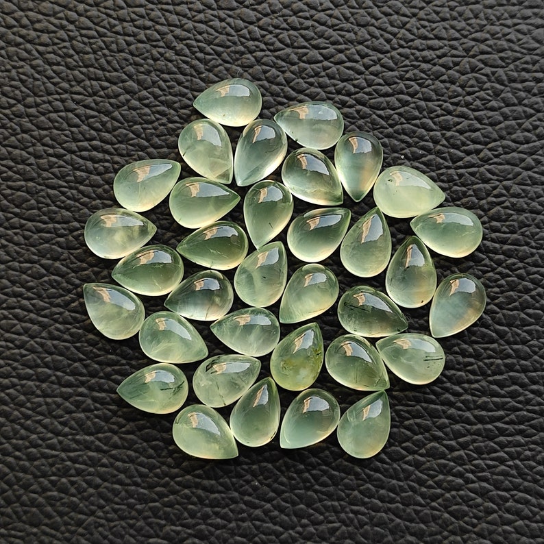 May include: A collection of 25 teardrop-shaped green gemstones. The stones are translucent and have a slight shimmer. They are arranged in a circular pattern on a dark background.