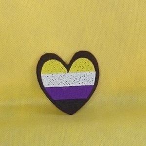 May include: A yellow, white, purple, and black heart-shaped patch with a stitched design. The patch is on a yellow background.