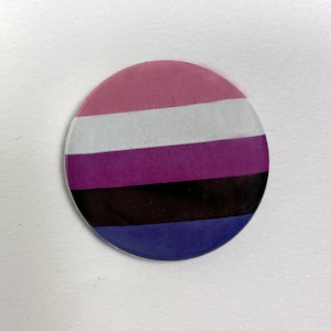 May include: A round button featuring a pink, white, purple, black, and blue striped design. The button represents the genderqueer pride flag.