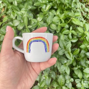 Può includere: Una tazza da caffè in ceramica bianca con un design arcobaleno dipinto a mano. L'arcobaleno è nei colori rosso, arancione, giallo, verde, blu, indaco e viola.