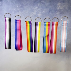 May include: Eight colourful keychains with different pride flag designs. The keychains are made of ribbon and have silver metal rings.