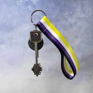 Może przedstawiać: Brelok do kluczy ze srebrnym kluczem i wstążką w kolorze fioletowym, białym, żółtym i czarnym.